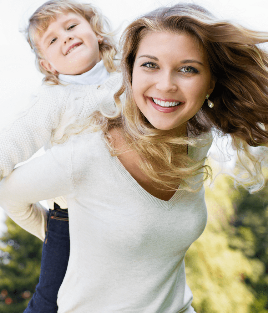 A woman playing piggyback with her child. Both are smiling during a sunny day.