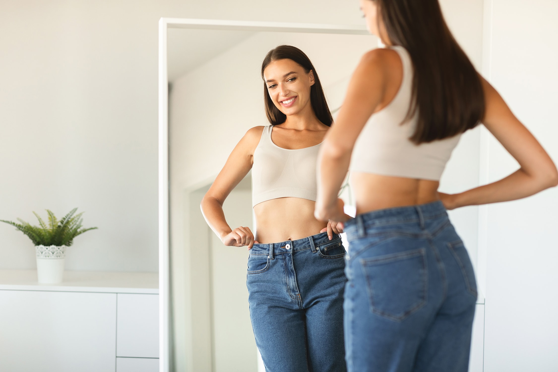 Cheerful young woman posing wearing jeans and smiling to her reflection in mirror after successful weight loss, copy space. Female beauty and style, self confidence, slimming concept