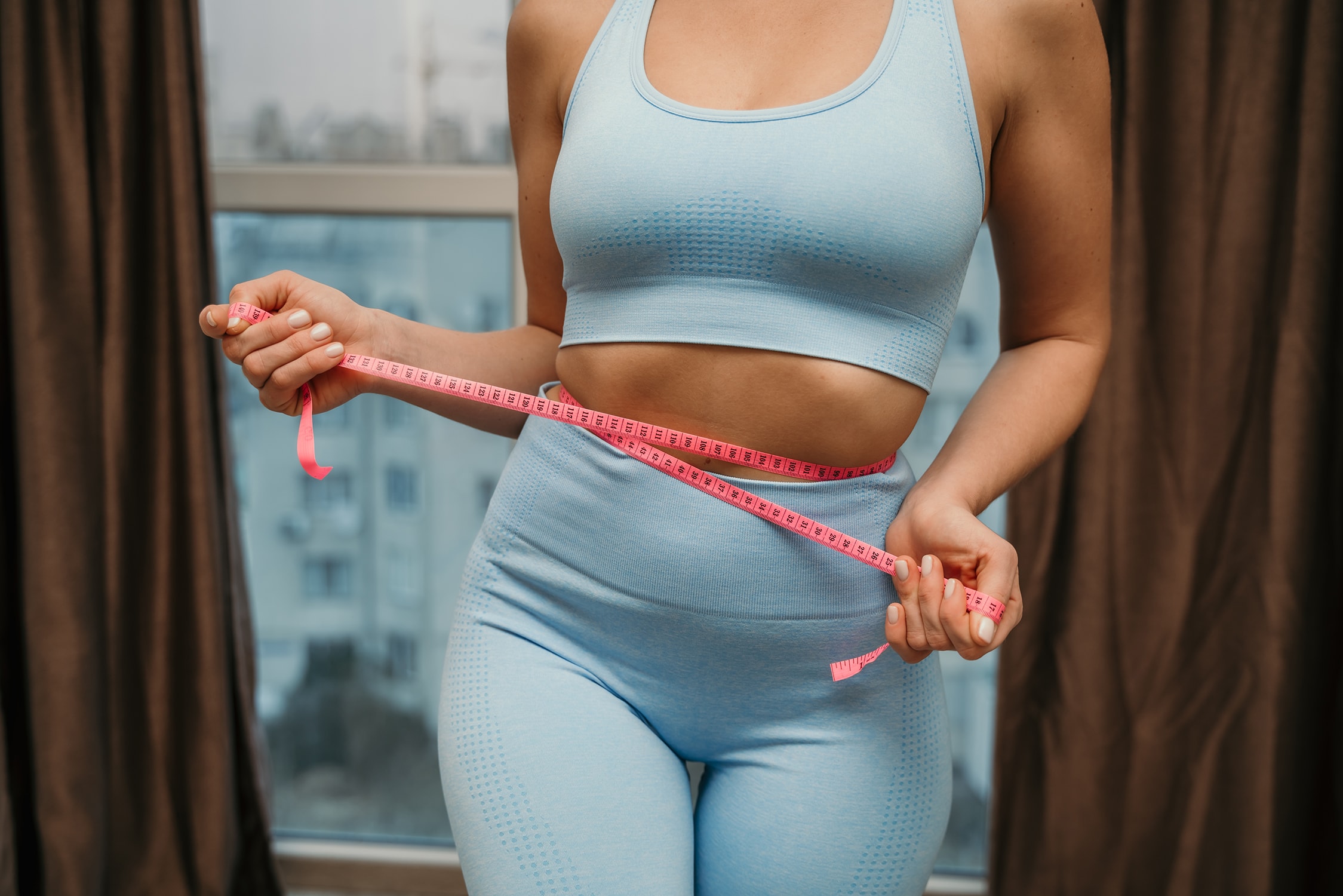 Cropped view of slim woman measuring waist with tape measure at home, close up. Unrecognizable european woman checking the result of diet for weight loss or liposuction indoors