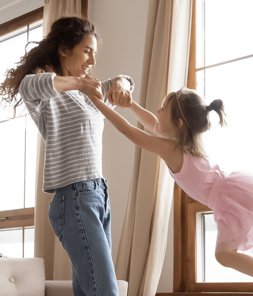 A woman playing with her daughter by spinning her around in the air. The daughter is wearing a pink tutu, and both are smiling and laughing in a sunny living room.